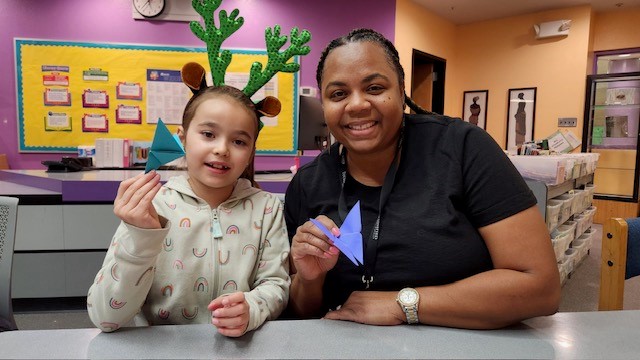 A student in green reindeer antlers and Principal Thomas smile and hold up their paper crafts.
